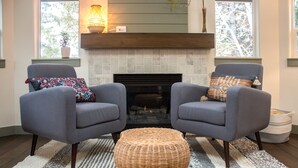 A living room with two grey armchairs, a wicker ottoman, a fireplace, and a striped rug. - Modern Midtown Home w/ Luxury Finishes & Hot Tub! (Bend)