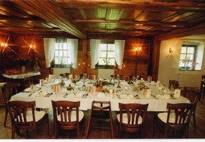 Uma sala de jantar com uma mesa comprida posta para uma refeição, paredes de madeira e janelas com cortinas.