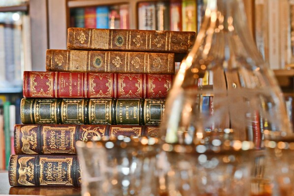 Una pila de libros antiguos con cubiertas ornamentadas, un florero de vidrio transparente y un fondo desenfocado de estanterías.