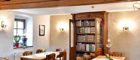 A room with wooden ceiling beams, a large open bookshelf, and a dining area with tables and chairs.