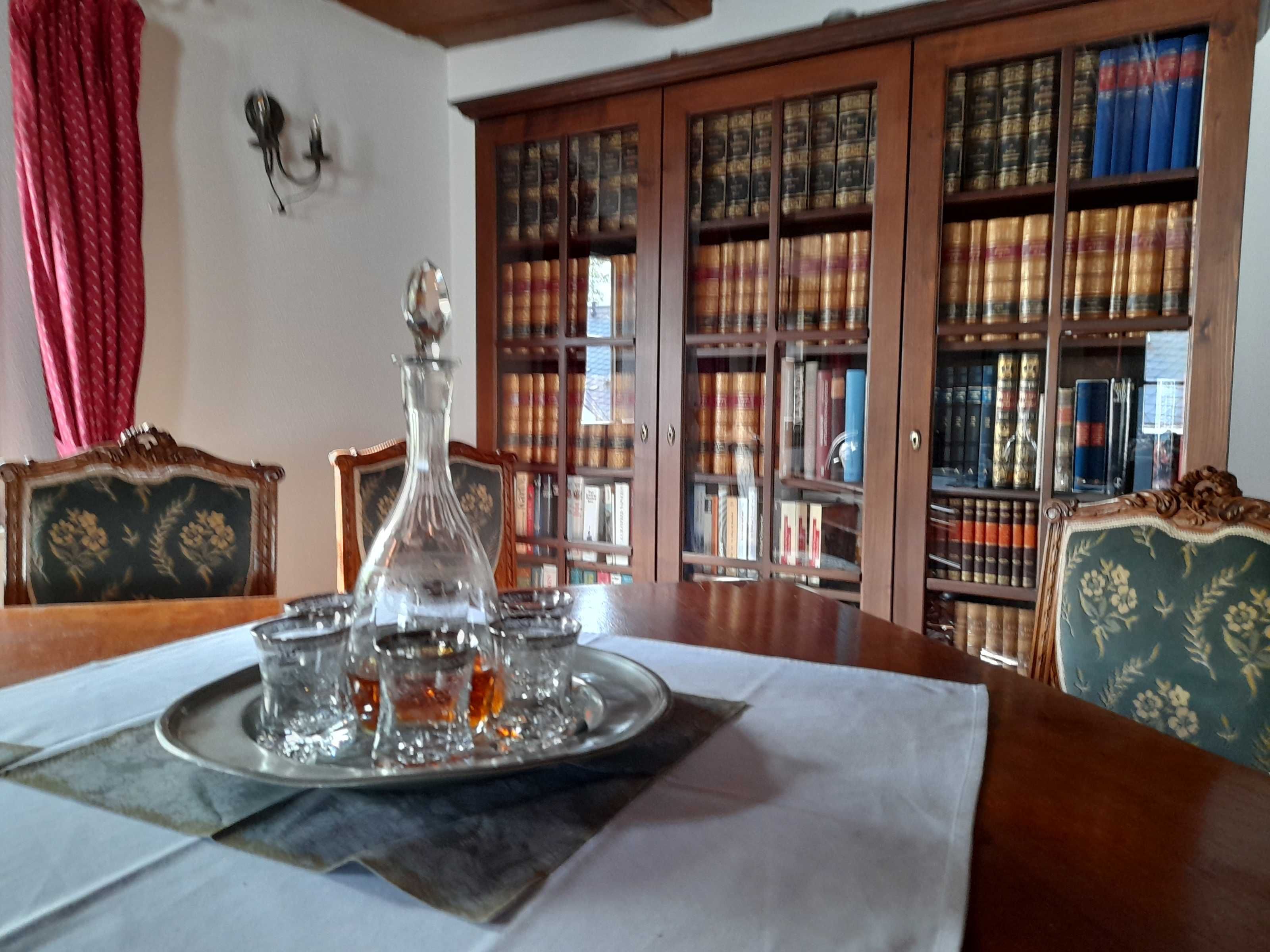 A room with a wooden table, a glass decanter, and a tray with glasses. There is a bookshelf filled with books in the background.