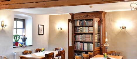 A room with wooden ceiling beams, a large open bookshelf, and a dining area with tables and chairs.