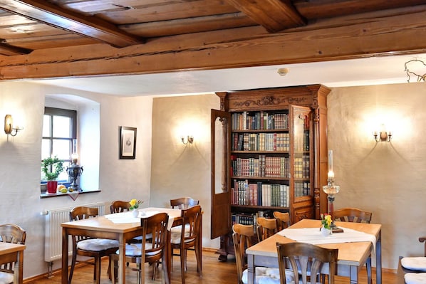 A room with wooden ceiling beams, a large open bookshelf, and a dining area with tables and chairs.