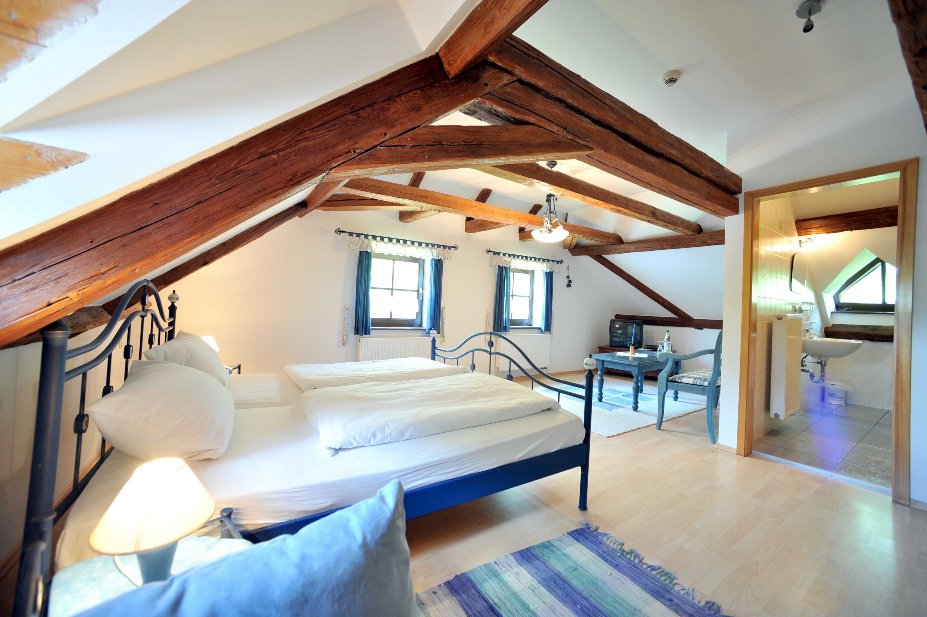 Ein Schlafzimmer mit Bett, Nachttischlampe, Holzbalken, einem Fenster und einem Badezimmer mit Waschbecken.