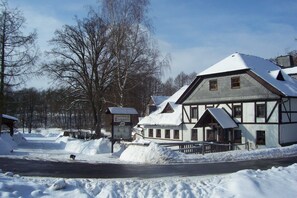 En snötäckt byggnad med en skylt som visar texten ”Gastrop Teichmühle”, en asfalterad väg och kala träd i bakgrunden.