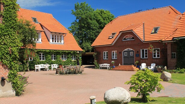 A courtyard with brick buildings, outdoor seating, and a small garden. - Der Besitz 6a (Stoltebuell)
