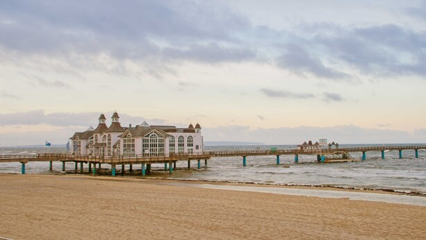 A coastal building with a pier extending into the sea, featuring a large bay window and a balcony.