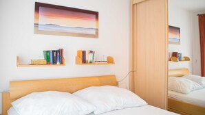 A hotel room with a wooden bed, white pillows, a bookshelf with books, a framed picture on the wall, and a mirror reflecting the room.