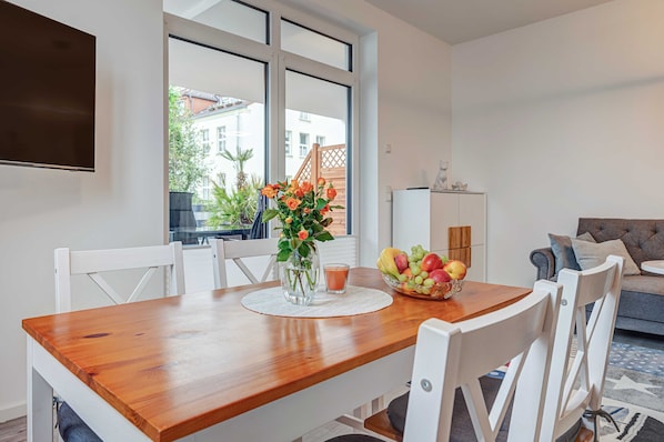 A dining area with a wooden table, white chairs, a vase with flowers, and a fruit bowl.