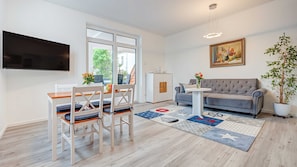A modern living room with a grey sofa, a dining table with chairs, a TV mounted on the wall, and a potted plant.
