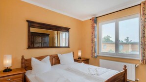 A neatly made bed with white linens, a wooden headboard, and two bedside tables with lamps. A large mirror hangs above the bed, and a window with curtains is visible. - Vacation apartment at Krebssee Whg. Otter - Otter (Heringsdorf)