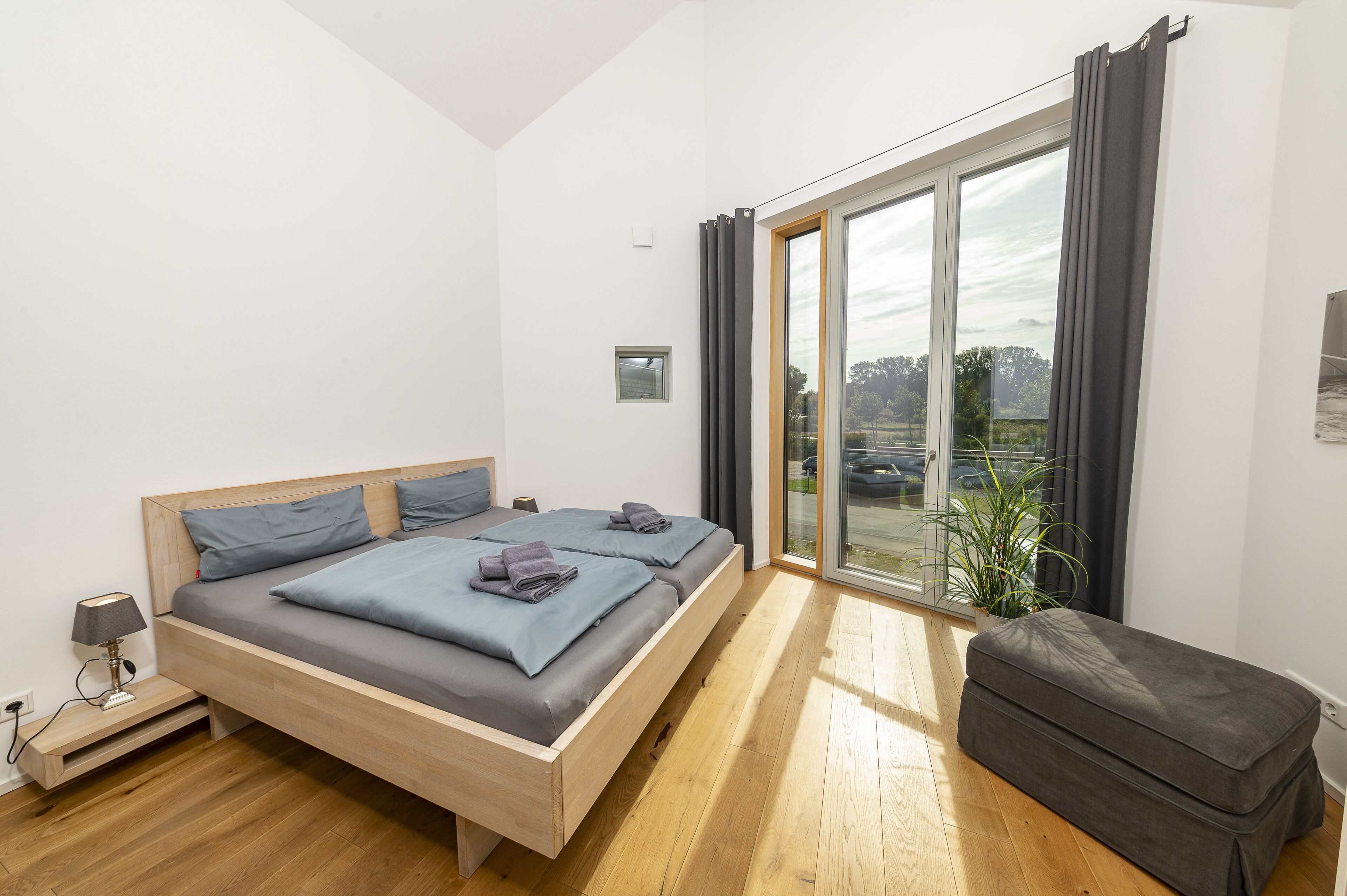 Un dormitorio moderno con una cama de madera, una mesita de noche pequeña, un banco gris y un ventanal con cortinas.