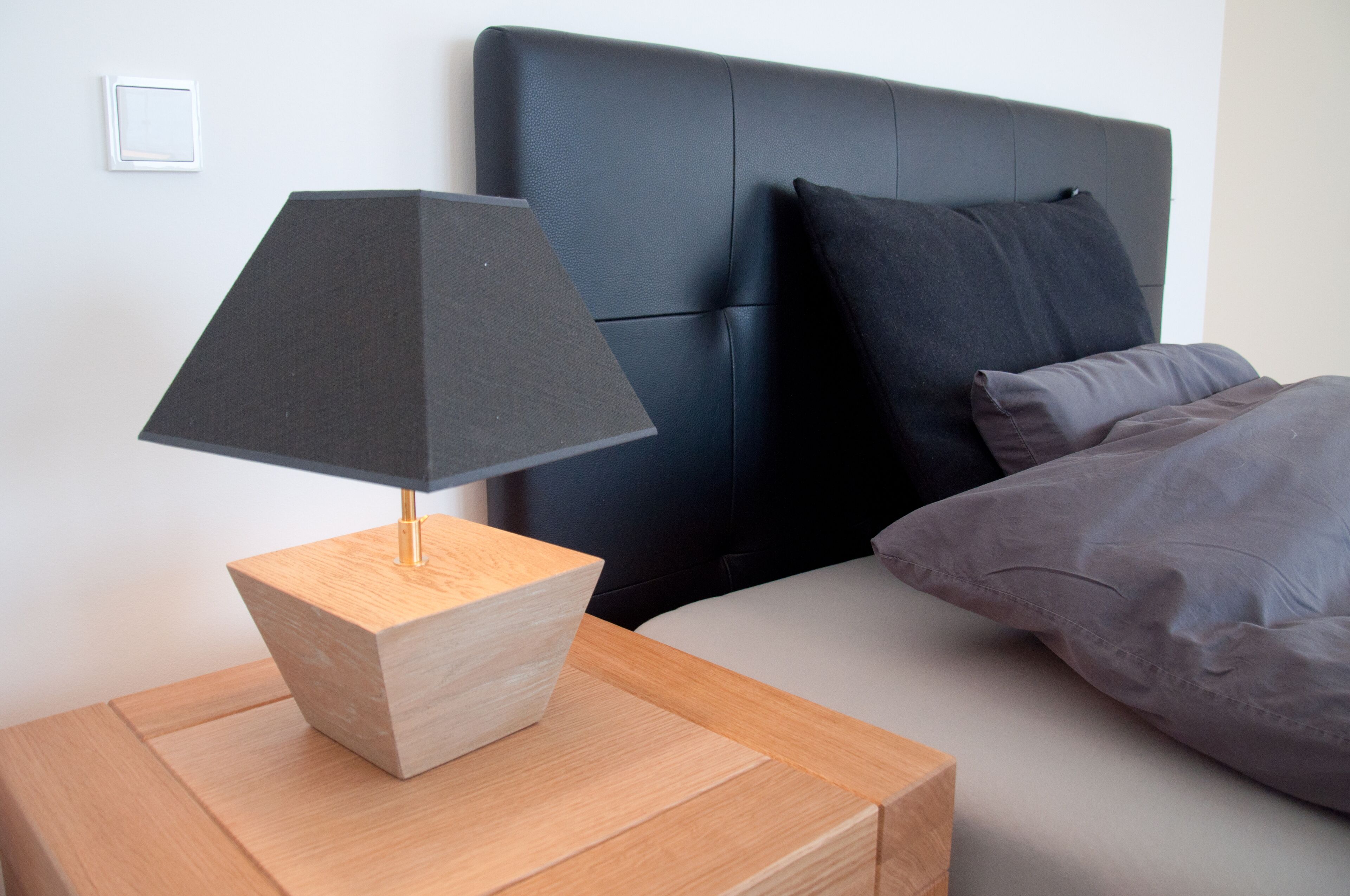 A modern bedroom with a wooden nightstand, a black lamp, a bed with grey bedding, and a dark headboard.