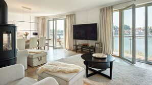 A modern living room with a large TV, comfortable seating, a fireplace, and a view of a body of water through large windows. - Kapitänsbrücke - Penthouse (Kappeln)