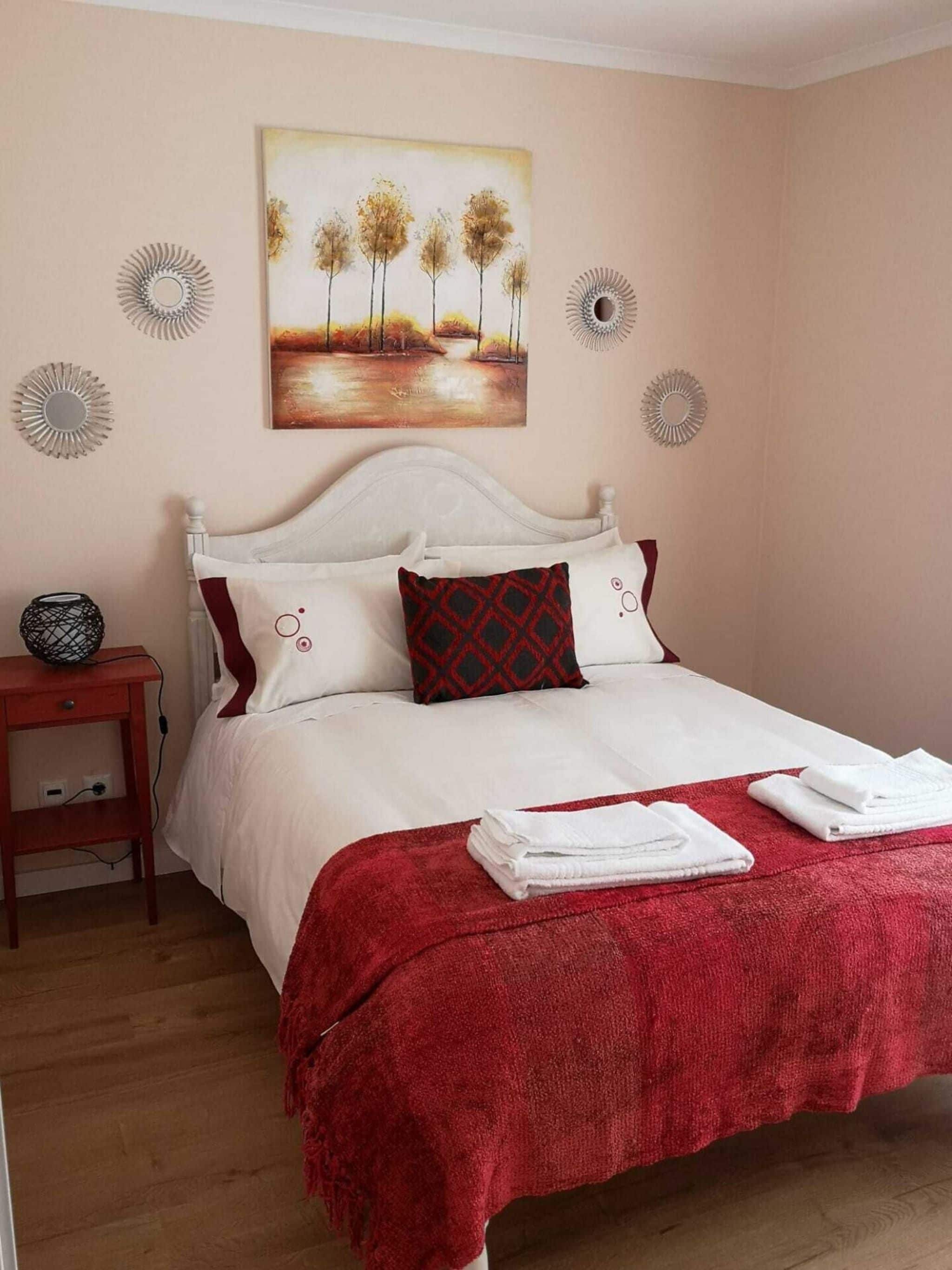A neatly made bed with white bedding and a red throw blanket, a painting of trees on the wall, and a small bedside table with a lamp.