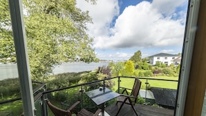 Ein Balkon mit Tisch und Stühlen mit Blick auf einen See und einen gepflegten Garten, im Hintergrund ein Haus.