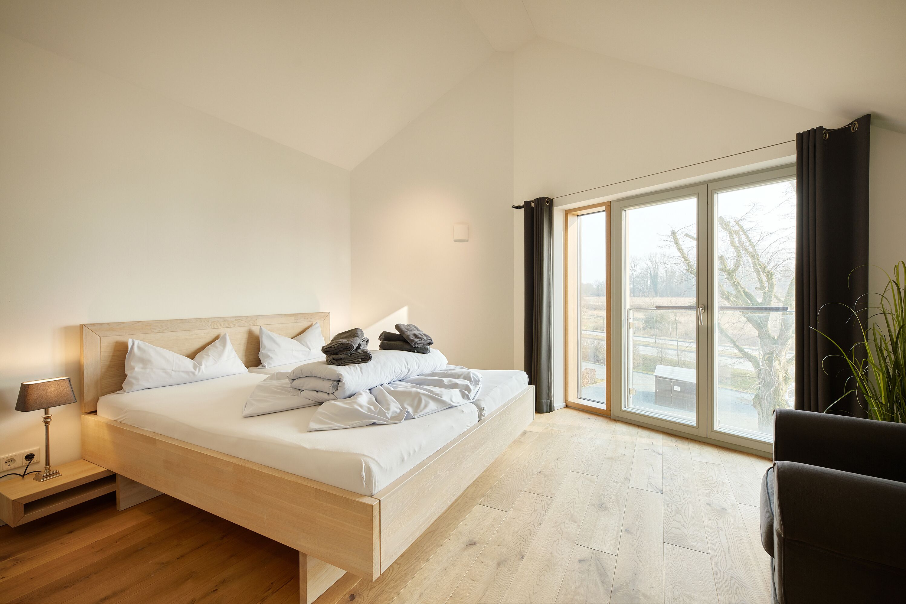 Un dormitorio moderno con una cama de madera, una mesita de noche con lámpara y un ventanal con vistas al exterior.