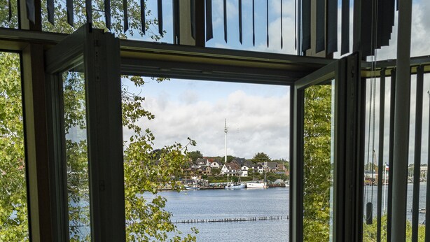 Une pièce dotée d’une grande fenêtre donnant sur un lac, d’un balcon avec une balustrade et d’une vue sur les maisons situées de l’autre côté de l’eau.