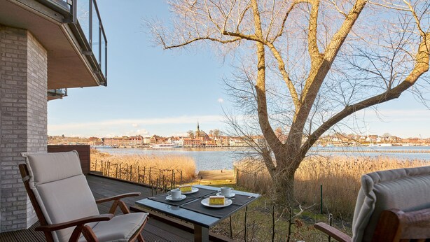 Une terrasse avec une table dressée pour deux personnes, offrant une vue sur un plan d’eau et sur le paysage urbain.