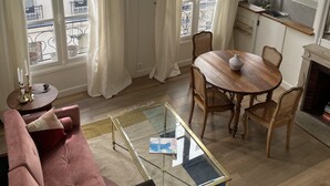 A living room with a sofa, a glass coffee table, a dining table with chairs, a fireplace, and a kitchen area with cabinets and a window.