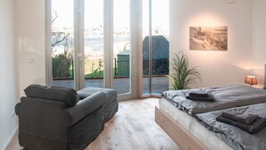 A modern bedroom with a large sliding glass door leading to a balcony, a bed with neatly folded towels, a gray sofa, and a wall-mounted artwork. - Steuermann (Kappeln)