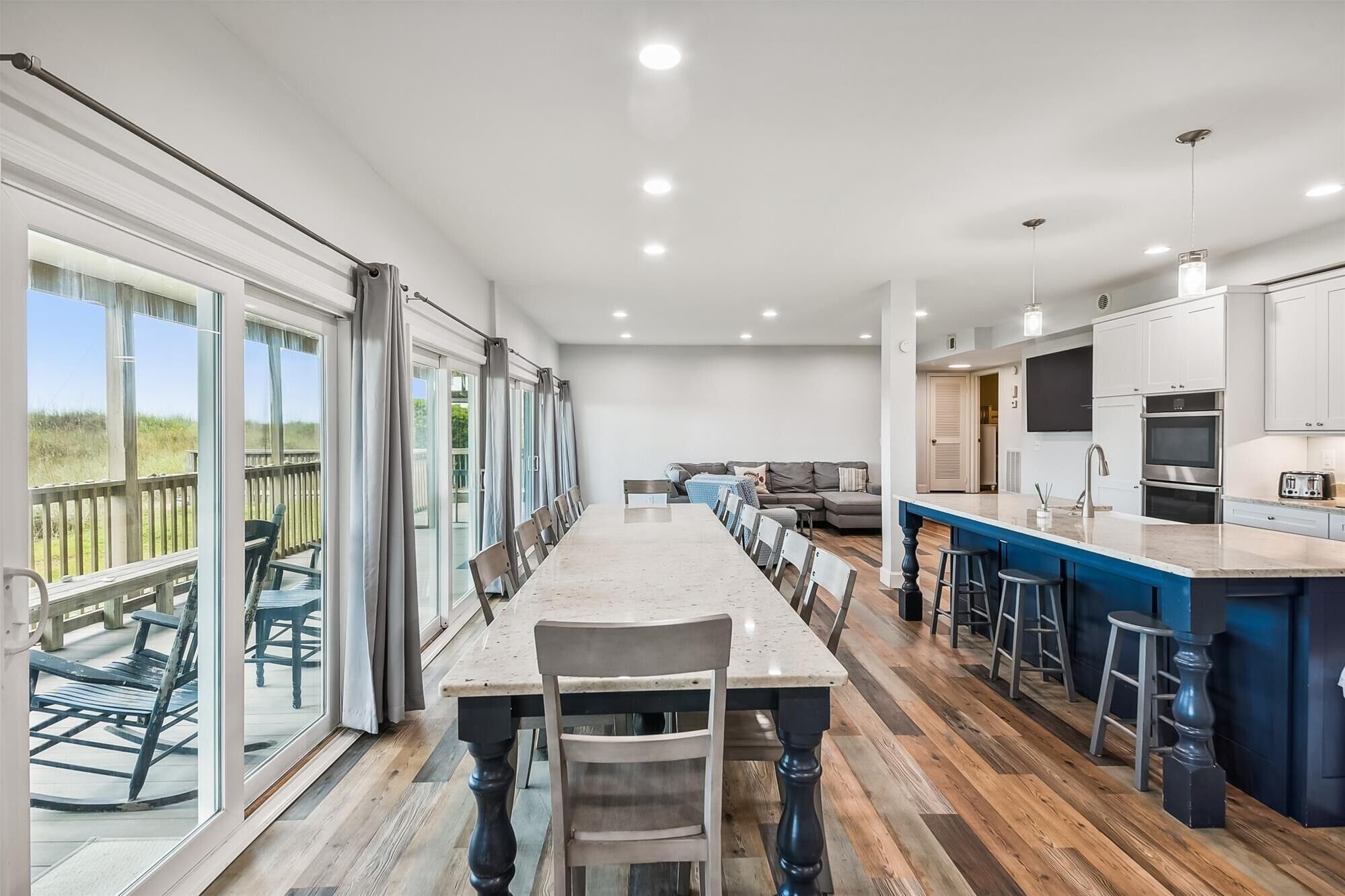 A modern kitchen with a large island, a dining area with a long table, and a view of the outdoors through large sliding glass doors.
