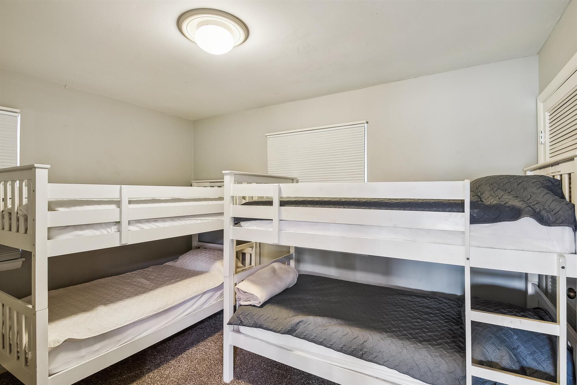 Een slaapkamer met een stapelbed, een plafondlamp, witte muren en een raam met jaloezieën.
