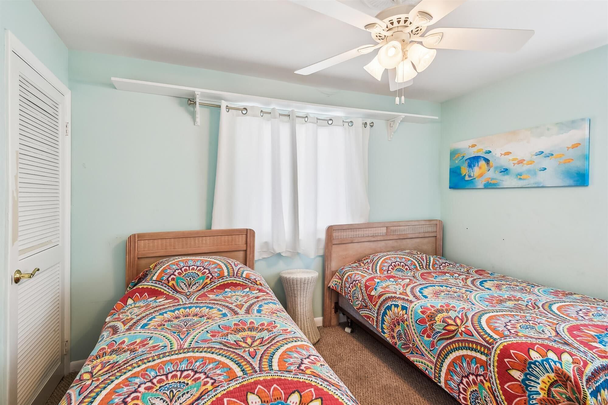 Une chambre avec deux lits simples, un ventilateur de plafond, une fenêtre avec des rideaux blancs et un tableau au mur.