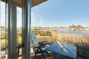 A balcony with a table and chairs, overlooking a river and a town with buildings and a church. - Lotsenhaus 12 (Kappeln)