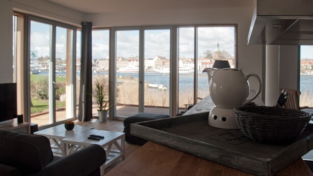 Un moderno salón con un amplio ventanal que da a un río y edificios. Hay un hervidor blanco, una cesta de mimbre y una mesa de madera.