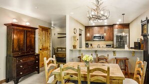A dining area with a wooden table and chairs, a chandelier, and a kitchen with wooden cabinets and modern appliances. - Ski OR Walk to Lifts, Village and Hot Tub! Luxurious & Clean Beaver Creek Condo (Beaver Creek)