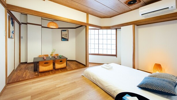 A room with a wooden ceiling, a bed with pillows, a bedside table, a window, and a dining area with a table and chairs. - TORA HOTEL Taito D (Tokyo)