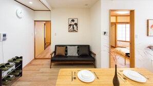 A room with a wooden dining table set for two, a black sofa with patterned cushions, a clock on the wall, and a traditional Japanese artwork. - TORA HOTEL Taito D (Tokyo)