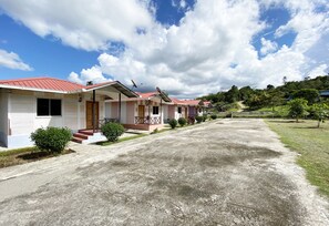 Una hilera de casas de un piso con tejados rojos y antenas parabólicas, un camino de grava y un paisaje montañoso al fondo.