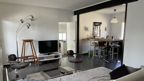 Ett modernt vardagsrum med en platt-TV, en soffa, en matplats med bord och stolar samt ett kök i bakgrunden.
