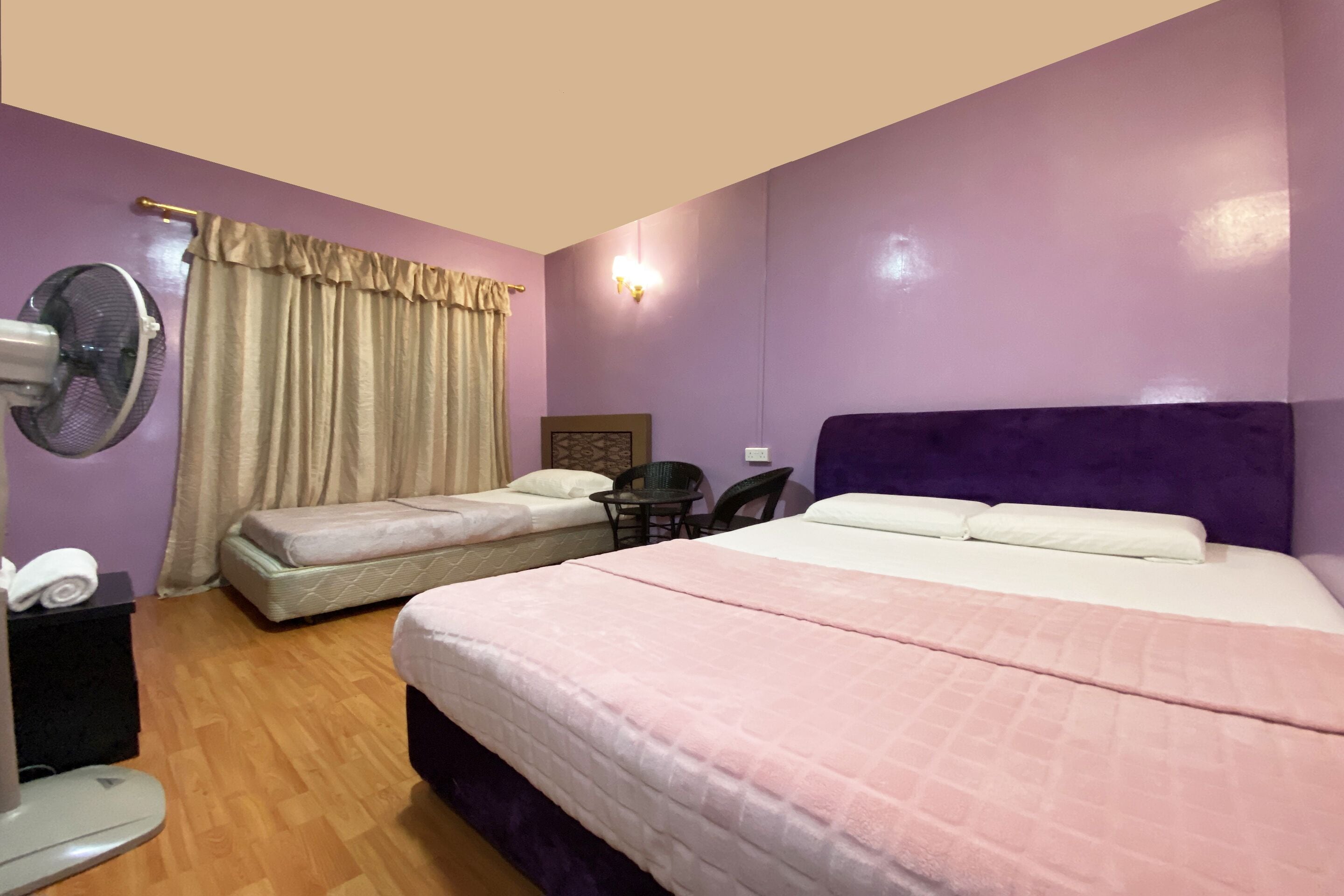 Une chambre d’hôtel avec deux lits, un ventilateur, un bureau avec une chaise et une fenêtre avec des rideaux.