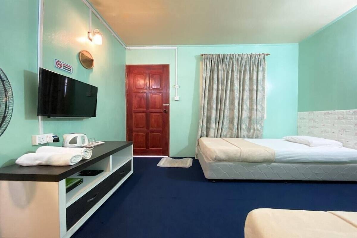 Habitación de hotel con cama, televisor montado en la pared, una pequeña mesa con tetera y ventilador, y una puerta que da a otra habitación.