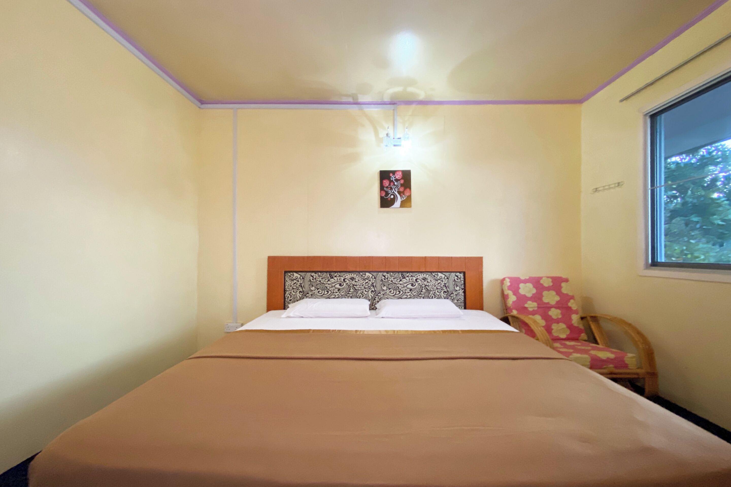 Una cama bien hecha con un cabecero estampado, una lámpara de pared sencilla y un ventanal con vistas a árboles.