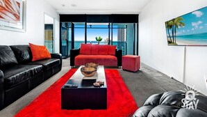 A modern living room with a black leather sofa, red cushions, a red rug, and a large window offering a view of the ocean. - Circle on Cavill – 1 Bedroom Unit with Ocean Views (Surfers Paradise)