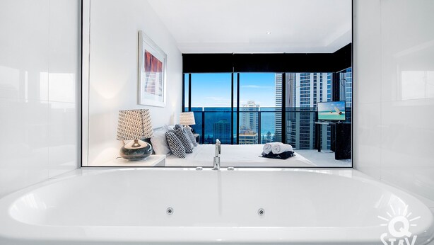 A modern bathroom with a large bathtub, a view of the cityscape through large windows, and a framed artwork on the wall. - Circle on Cavill – 1 Bedroom Unit with Ocean Views (Surfers Paradise)
