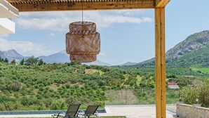 Una pérgola de madera con una lámpara colgante de mimbre, un patio de concreto con dos sillones de descanso y una vista de colinas y montañas verdes al fondo.