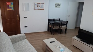Ein Wohnzimmer mit einer Couch, einem Couchtisch mit Fernbedienung und Schlüsseln, einem Fernseher auf einem Lowboard sowie einem Essbereich mit Tisch und Stühlen.