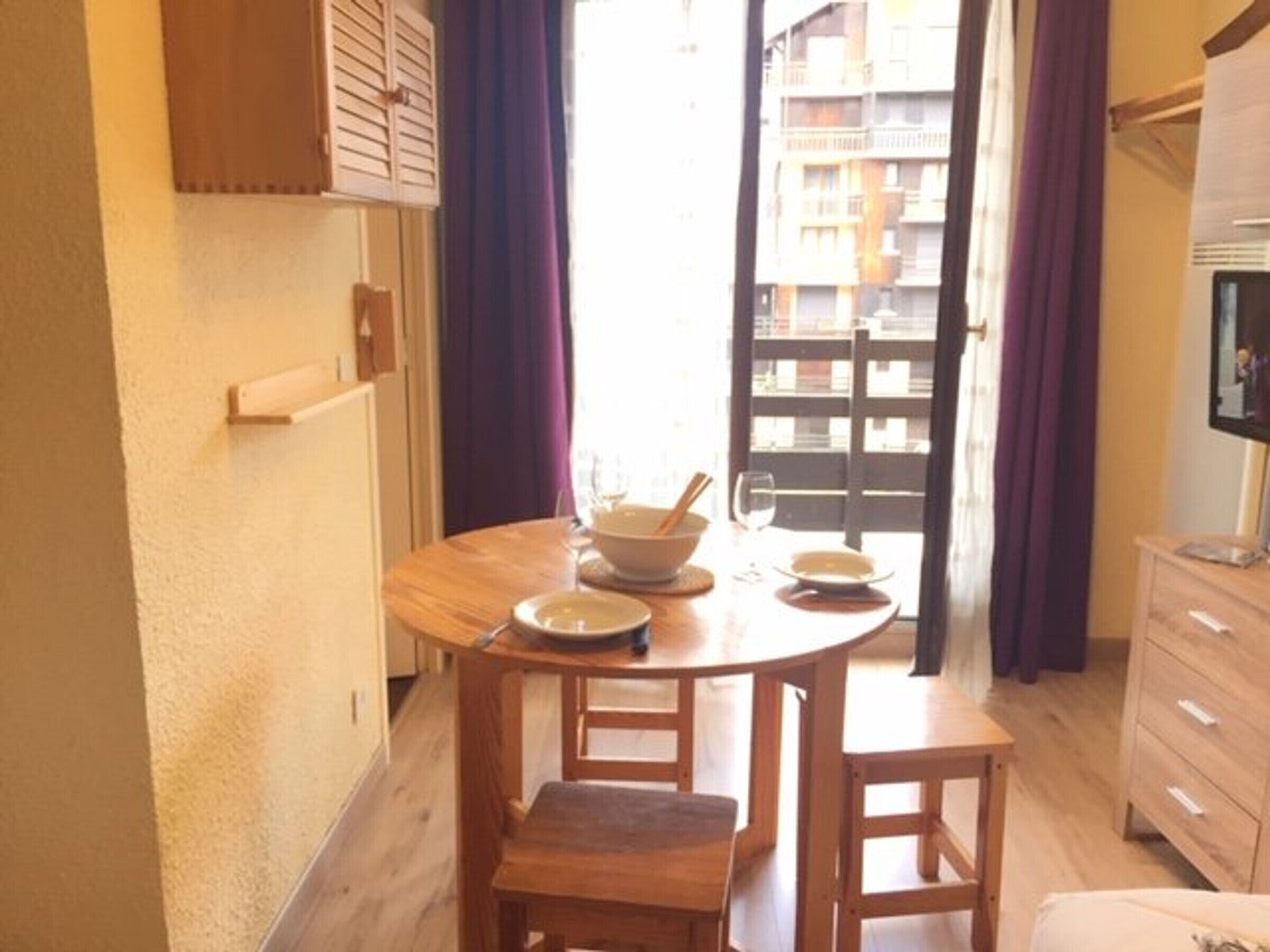 A small dining area with a round wooden table, two chairs, and a bowl with a spoon. There is a window with purple curtains, and a TV is mounted on the wall.