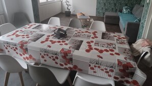 A dining area with a table covered in a floral tablecloth, surrounded by white chairs. A potted plant is on the windowsill, and a clock is on the wall. - Logement Entièrement Neuf Tout Confort (Lucey)