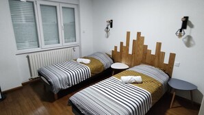 A bedroom with two beds, a radiator, a standing heater, a window with blinds, and a small round table. - Logement Entièrement Neuf Tout Confort (Lucey)