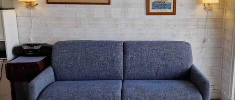 Un canapé bleu dans une pièce aux murs en briques blanches, deux tableaux encadrés et une petite table sur laquelle se trouve une radio.