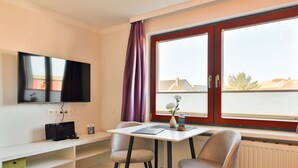 Ein Zimmer mit an der Wand befestigtem Fernseher, Tisch mit Stühlen, einem Fenster mit Vorhängen und einem Heizkörper darunter.