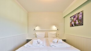 Cama de casal com lençóis e toalhas brancas, cabeceira com ripas horizontais e luminária fixada na parede.