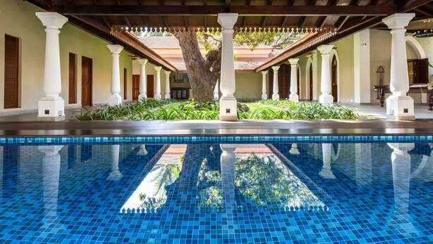Une piscine dotée d’un sol carrelé et de colonnes blanches, entourée de verdure et agrémentée d’un espace couvert avec un toit carrelé.