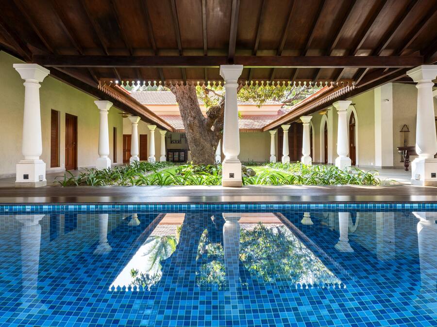 Piscina con suelo de baldosas y columnas blancas, rodeada de vegetación y una zona cubierta con techo de tejas.